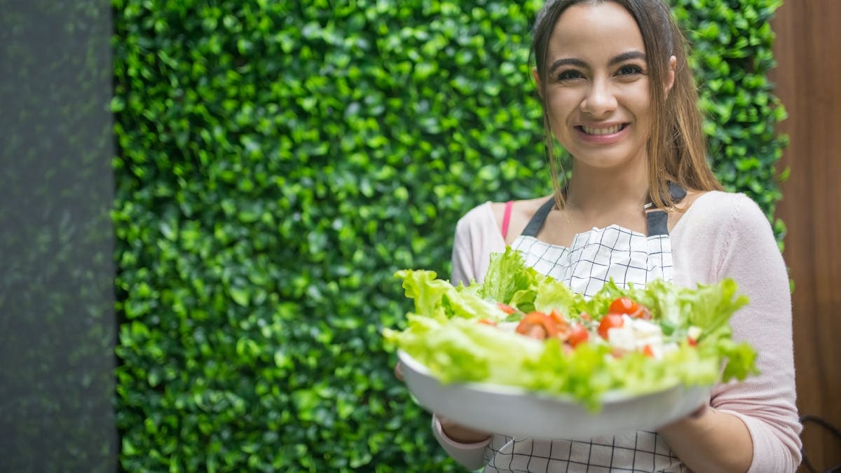 Woman in apron presenting a fresh vegetable salad. Perfect for healthy living and cooking themes.